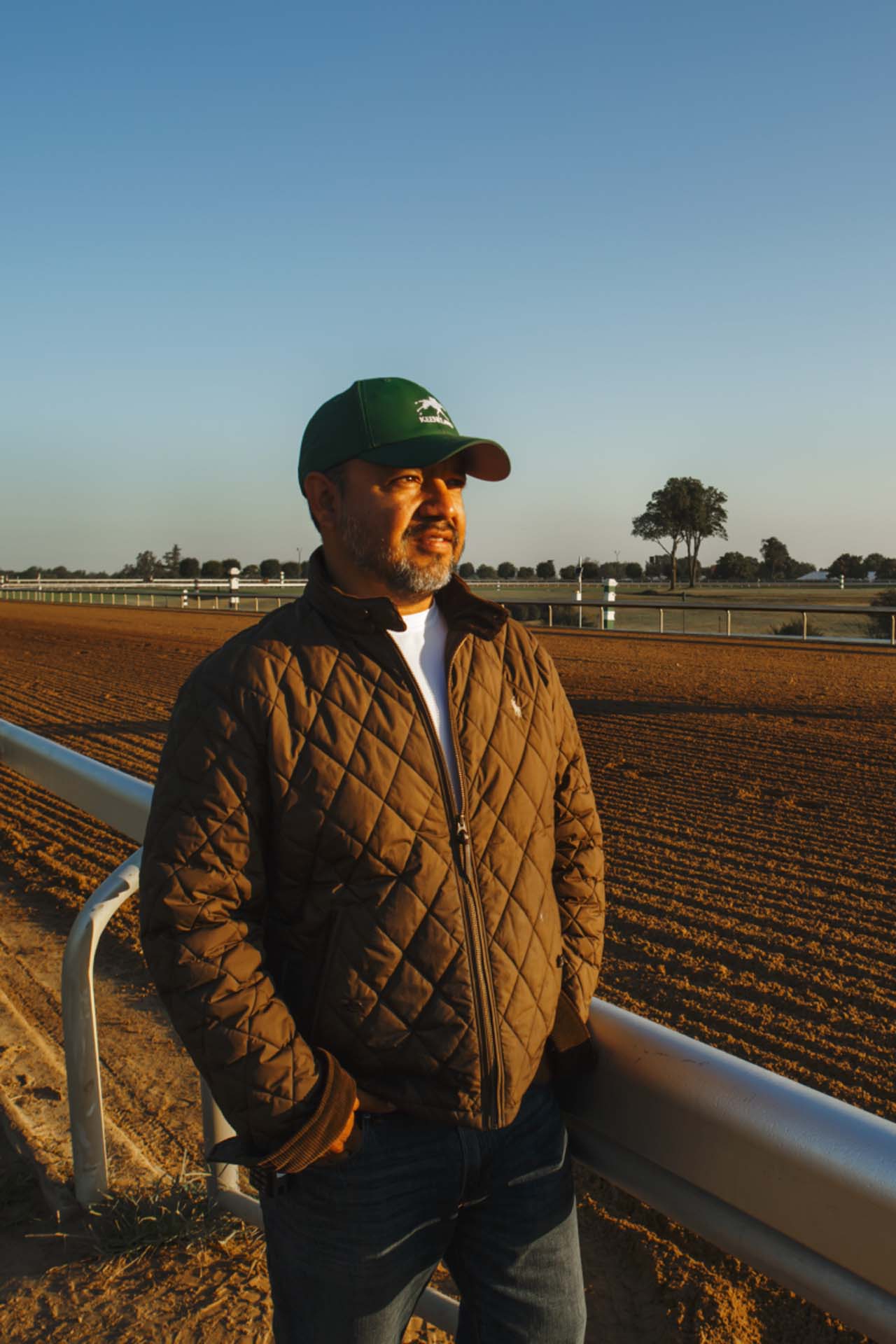 A photo of Alfredo Laureano looking off into the distance while standing by the dirt track at sunset. He is an older Hispanic man with graying chin stubble. He is wearing a green Swiftjockey-brand hat and a brown Swiftjockey-brand jacket overtop a white shirt.