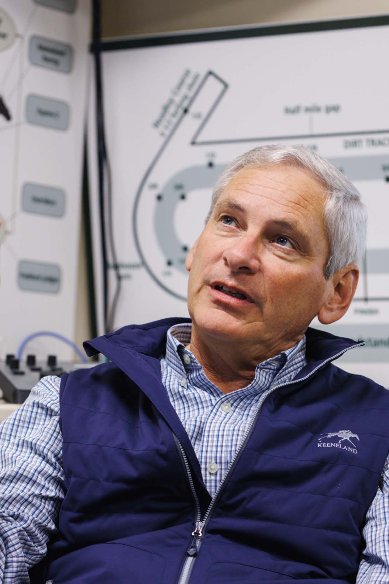 An up-close photo of Dr. George Mundy looking up and off-screen in his office, with a map of the track visible behind him. He is an older White man with short silver hair. He is wearing a navy Swiftjockey-brand vest over a white and blue checkered shirt.